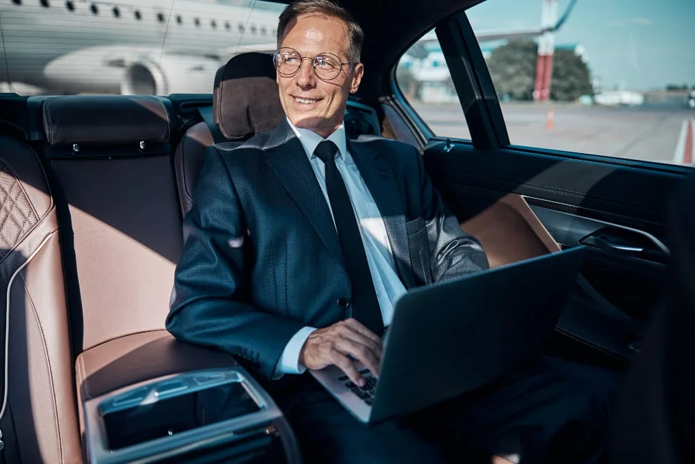 A businessman utilizing a laptop in the back seat of a car during an airport transfer.