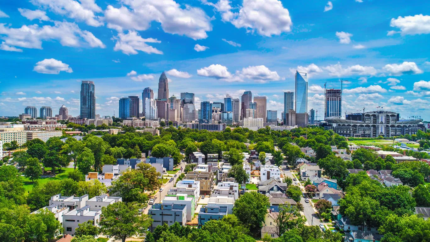 Aerial view of a city skyline with tall buildings, a stadium, and residential areas surrounded by trees under a blue sky with scattered clouds.