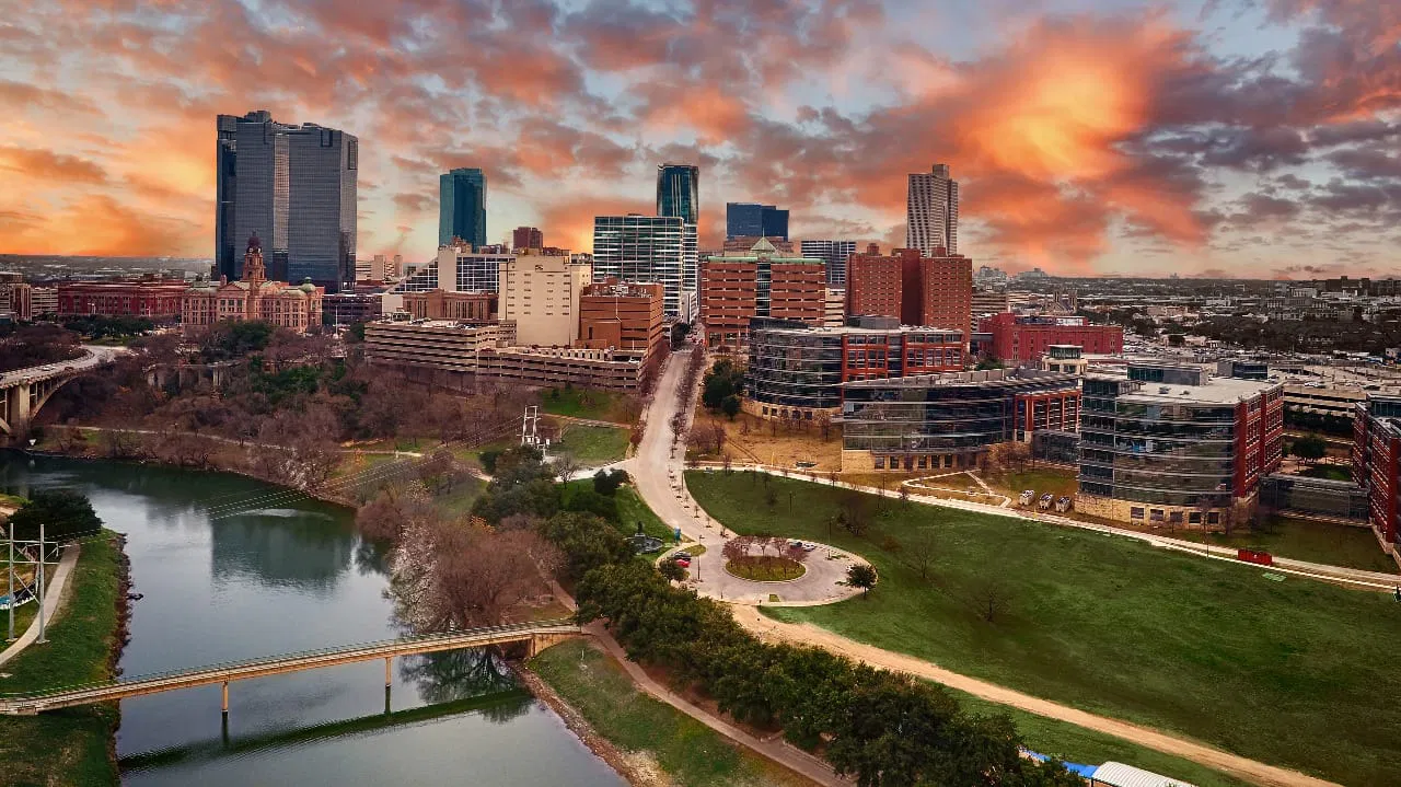 Aerial view of a cityscape featuring tall buildings, a river, bridges, and green spaces under a vibrant sunset sky.