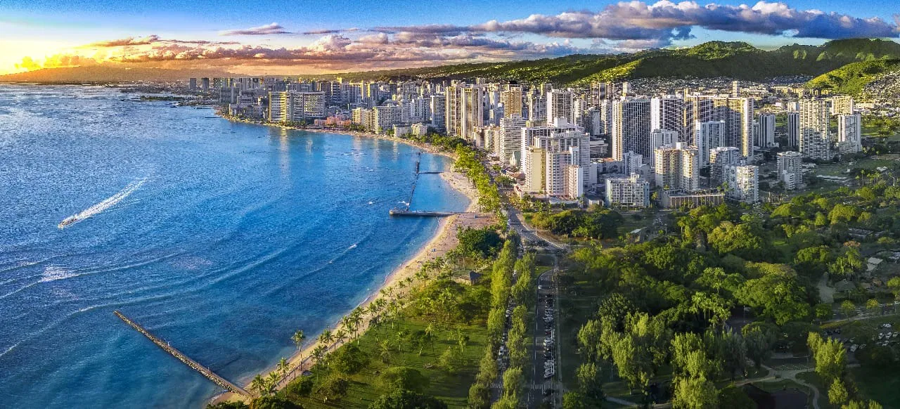 Aerial view of a coastal city with high-rise buildings along the waterfront, a sandy beach, ocean waves, and a mountain range in the background at sunset.