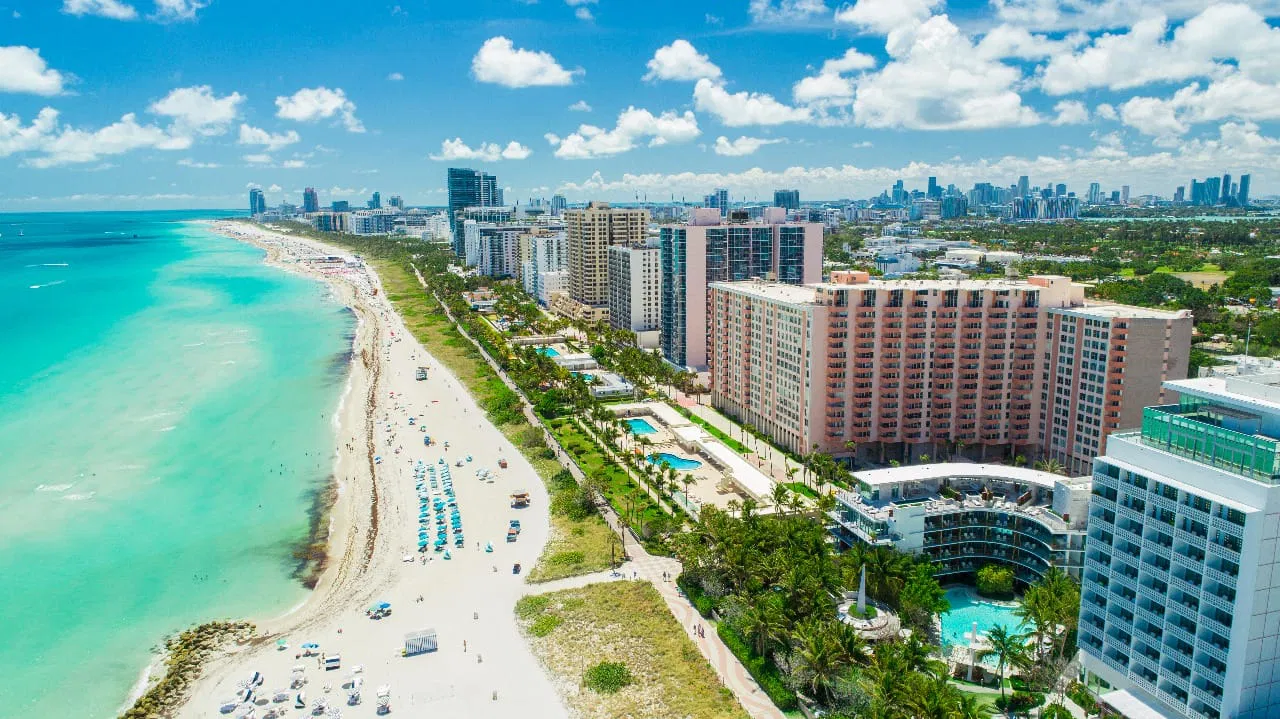 Aerial view of a beach with turquoise waters and sandy shores lined with umbrellas and sunbathers, adjacent to a cityscape of high-rise buildings and palm trees under a clear blue sky.