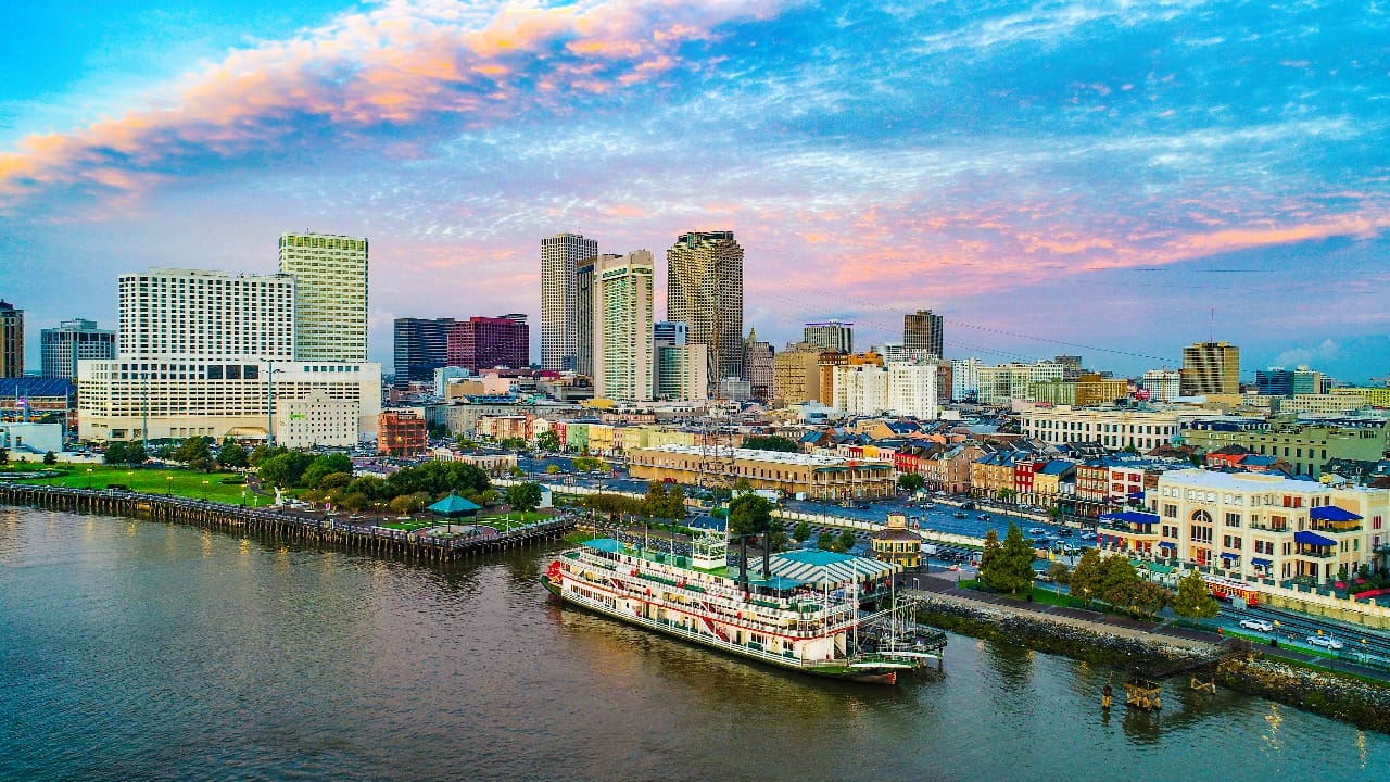 A riverboat on calm water near a bustling city skyline at sunrise, with a mix of modern skyscrapers and historic buildings under a partly cloudy sky.