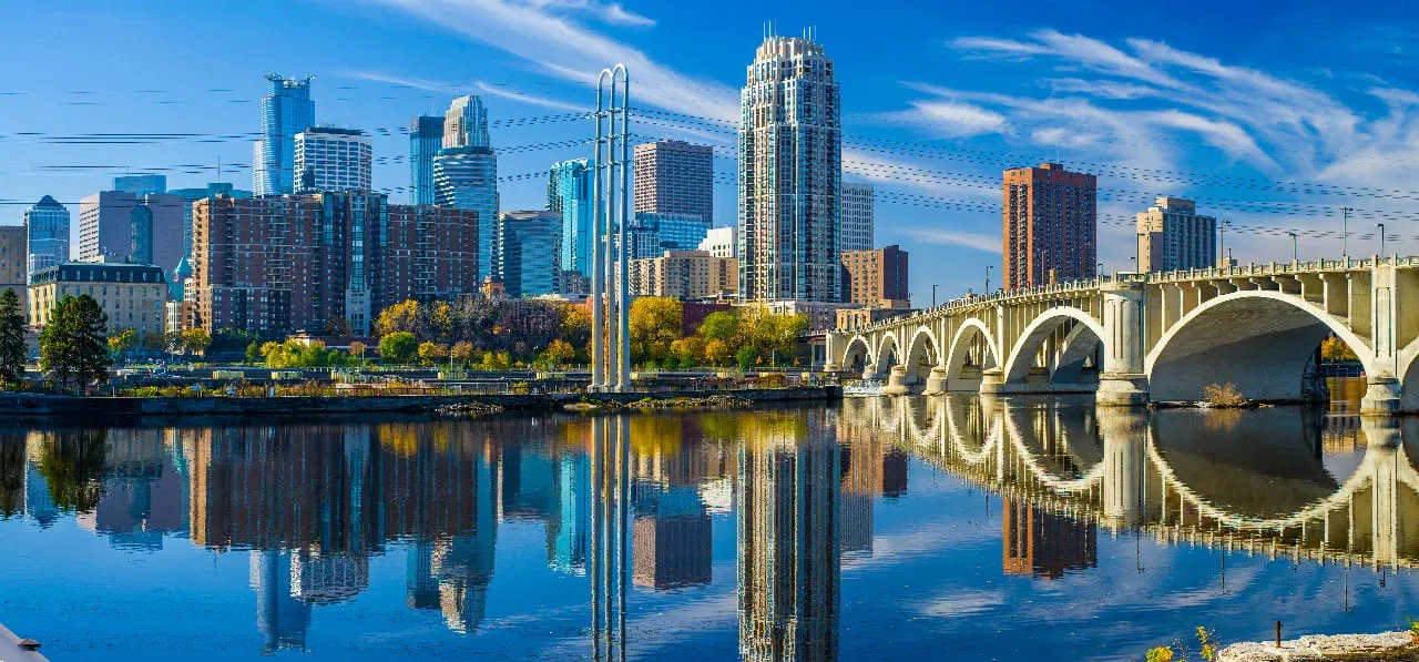 A city skyline with numerous skyscrapers is reflected in a calm river. An arched bridge spans the water, and the sky is clear and blue with some light clouds.