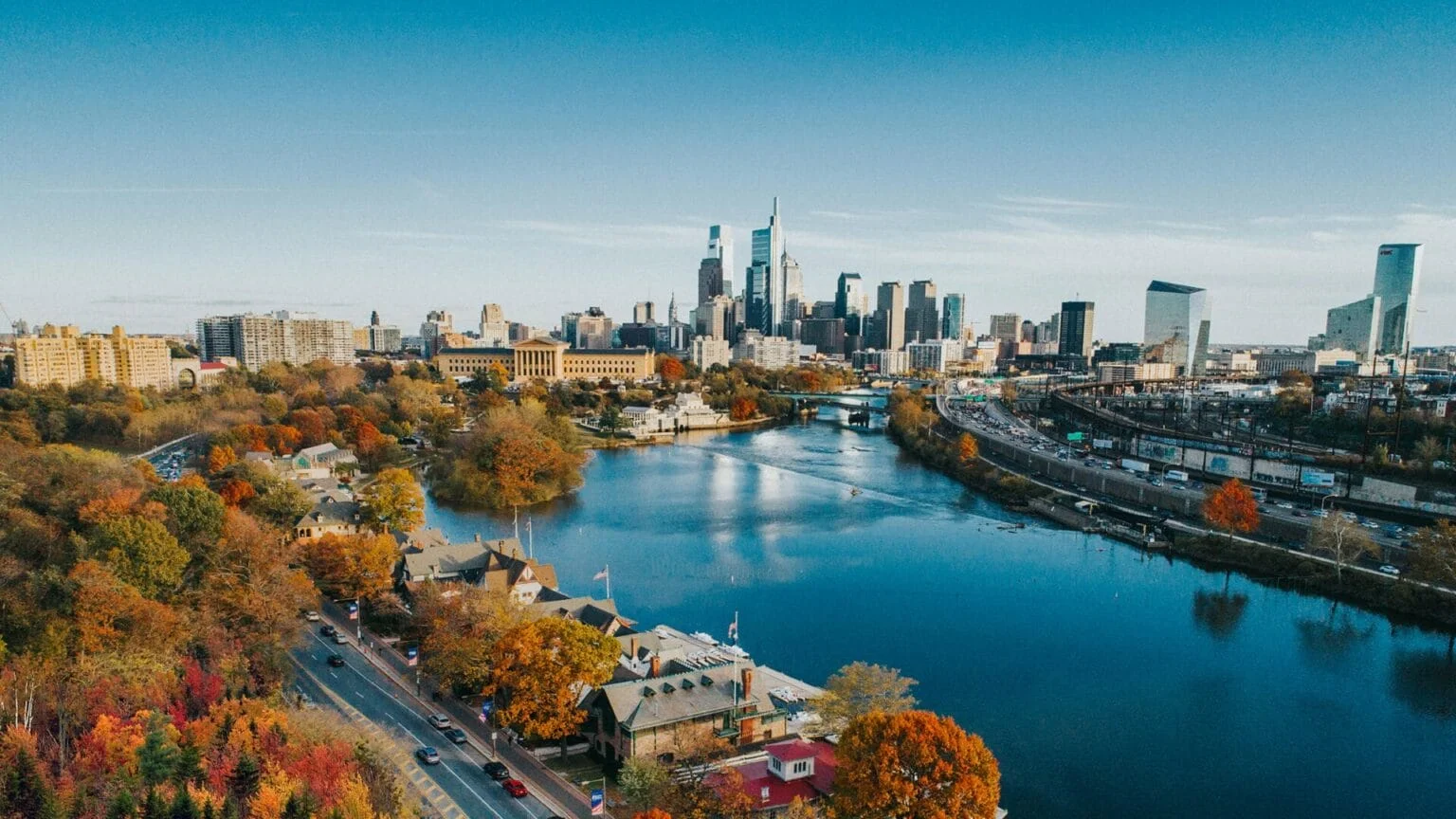 Aerial view of a river with trees showcasing fall colors, buildings, and a city skyline in the background under a clear blue sky.